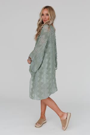 A woman with long blonde hair wears the Bali Embroidered Kimono in sage, a lightweight textured dress, and beige wedge sandals. She stands against a white background, smiling and looking over her shoulder.
