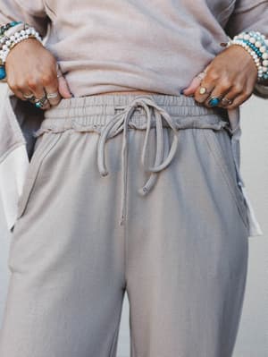 A close-up shot focused on the smocked drawstring waistband of soft ash-toned sweatpants, styled with a matching top and layered boho jewelry — the kind of outfit that makes staying in feel intentional.