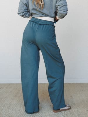 Shot from the back, a model shows off wide-leg sweatpants in a muted teal-jade, paired with a washed gray cropped sweatshirt — the kind of outfit that makes staying home feel like a whole personality.