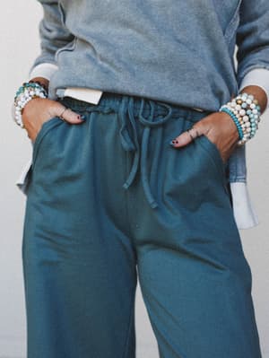 A close mid-shot of wide-leg sweatpants in a muted teal-jade tone, hands tucked into front pockets and stacked beaded bracelets adding a personal, collected touch against a neutral studio backdrop.