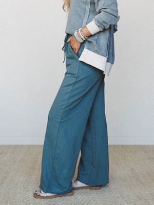 A side-profile shot of wide-leg sweatpants in a muted teal-jade tone, styled with a washed blue cropped sweatshirt and layered beaded bracelets — the kind of outfit that looks like you tried just the right amount.