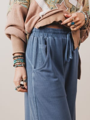 Person wearing a brown jacket, blue pants, and multiple bracelets on a neutral background