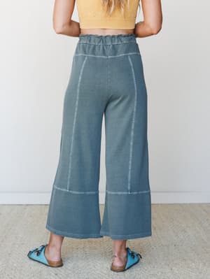 A back-view shot of wide-leg cropped lounge pants in a washed ash jade colorway, styled with a yellow crop top and turquoise footbed sandals against a clean studio wall — relaxed, lived-in, and effortlessly put-together.
