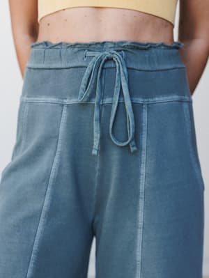 A close-up detail shot of wide-leg lounge pants in a washed ash jade blue, styled with a cropped yellow ribbed top — the drawstring waist and frayed waistband edge are the star of the show here.