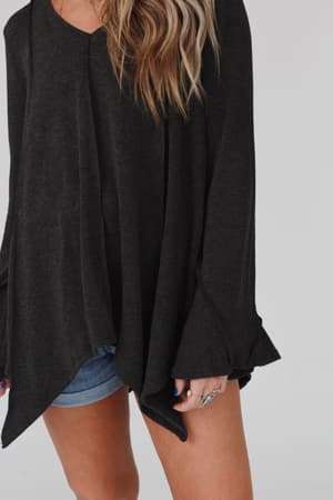 Someone stands against a plain light background wearing the Golden Hour Long Bell Sleeve Top in Charcoal with blue denim shorts. The loose-fitting, long bell sleeves add style, and their face is not visible.
