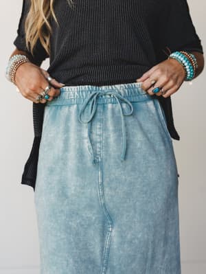 A close-up mid-shot of a mineral-washed maxi skirt in faded ash blue, styled with a dark charcoal ribbed top and layered boho jewelry — the kind of effortless pairing that feels thought-out without trying.