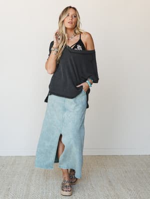 A relaxed full-body studio shot — acid-wash denim maxi skirt paired with an off-shoulder striped top, layered boho jewelry, and studded platform sandals, giving off that effortless 'ran into her at the farmers market and immediately texted my friends