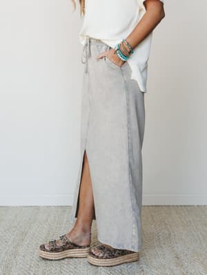A side-profile shot of a mineral-washed maxi skirt in a soft slate gray, styled with a cream waffle knit top, stacked turquoise bracelets, and leopard print platform sandals — the kind of effortless outfit that looks like it came together without try