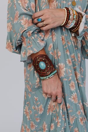 A person in a blue floral dress is shown from the neck down, wearing multiple rings and the Aegean Boho Leather Cuff Bracelet - Brown with earthy tones and turquoise accents on their wrist.
