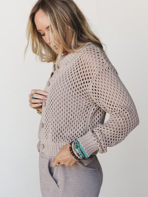 A cropped open-knit cardigan in warm ash mocha is styled with soft gray wide-leg pants, shot from the side as the model glances down — layered bracelets and stacked rings adding that effortlessly personal touch.