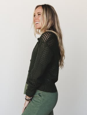 A smiling model shot from the side in a dark olive open-knit cardigan layered over an olive top with matching pants — the whole look reads effortless and earthy, like she just grabbed something cozy on her way out the door.