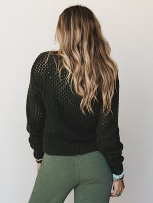 Shot from behind, a model with loose beachy waves wears a dark olive open-knit sweater paired with sage green leggings and stacked turquoise and pearl bracelets — effortlessly earthy and lived-in.