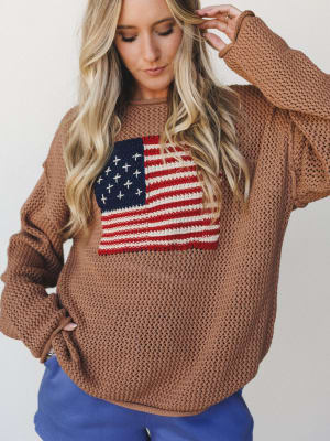 A relaxed open-knit sweater in deep camel with a bold American flag intarsia knit across the chest, styled with blue pants for an easy, expressive everyday look.