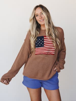 A relaxed open-knit sweater in deep camel with a bold American flag graphic on the chest, paired with periwinkle mini shorts — casual, easy, and just the right amount of festive without trying too hard.