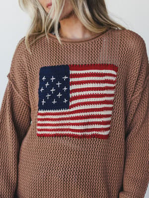 An open-knit sweater in deep camel with a chunky American flag intarsia knit front — the kind of piece that feels vintage-borrowed and intentionally worn, not costumey.