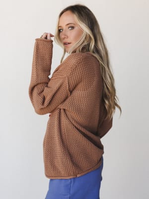 A model shot over the shoulder in a roomy open-knit sweater in deep camel — the kind of piece that looks equally at home on a coffee run or a cozy night in, paired here with a pop of periwinkle blue.