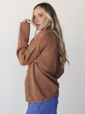 A model shot over the shoulder in a roomy open-knit sweater in deep camel — the kind of piece that looks equally at home on a coffee run or a cozy night in, paired here with a pop of periwinkle blue.