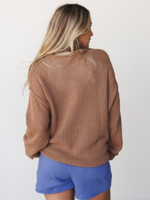 A relaxed open-knit sweater in warm camel is shown from the back, paired with periwinkle mini shorts — the kind of effortless combo that looks like you just threw it on and somehow nailed it.