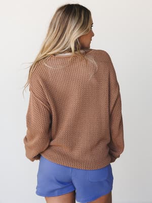 A relaxed open-knit sweater in warm camel is shown from the back, paired with periwinkle mini shorts — the kind of effortless combo that looks like you just threw it on and somehow nailed it.