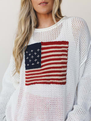 A relaxed open-knit ivory sweater with a hand-stitched American flag graphic on the chest — the kind of piece that makes a holiday weekend feel effortlessly intentional.