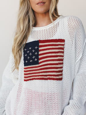 A relaxed open-knit ivory sweater with a hand-stitched American flag graphic on the chest — the kind of piece that makes a holiday weekend feel effortlessly intentional.