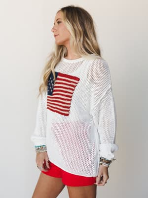 An oversized ivory open-knit sweater with a woven American flag graphic on the chest, styled over red mini shorts with stacked beaded bracelets — effortlessly patriotic and totally wearable.
