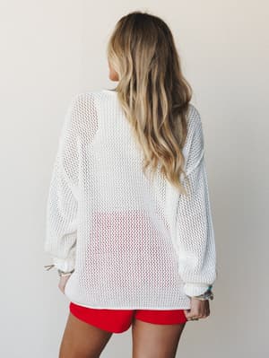 Shot from behind, a model wears an oversized open-knit ivory sweater over red mini shorts — the kind of effortless summer layering that looks like you grabbed it on the way out the door and somehow got it exactly right.