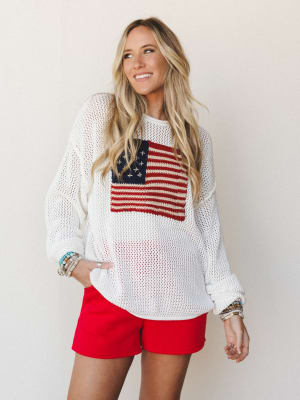 An oversized ivory open-knit sweater with a bold American flag intarsia graphic worn over red mini shorts — casual, sun-warmed, and ready for a backyard cookout with stacked bracelets on both wrists.