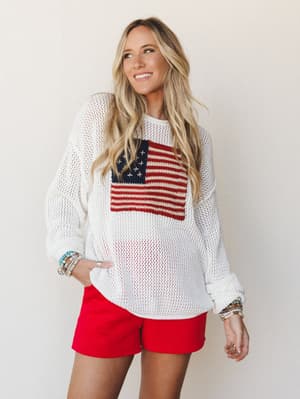 An oversized ivory open-knit sweater with a bold American flag intarsia graphic worn over red mini shorts — casual, sun-warmed, and ready for a backyard cookout with stacked bracelets on both wrists.