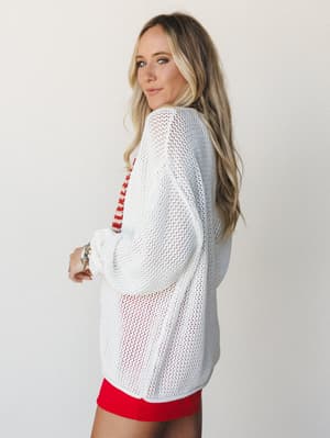 A relaxed open-knit ivory cardigan layered over a red striped top and red shorts — effortless Americana energy shot against a clean studio backdrop.