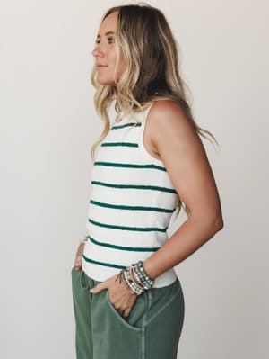 A side-profile studio shot of a model in a cream and hunter green striped sweater tank tucked into sage green relaxed pants, styled with layered beaded bracelets and a delicate necklace — effortless and easy, like she's always dressed this well.