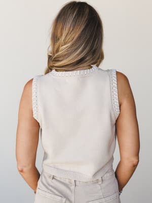 Shot from behind, this sand beige sweater tank has a quiet, understated charm — the contrast white whipstitch trim along the armholes and neckline does all the talking, paired with soft khaki pants for a tonal, easy look.