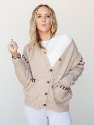 An oversized camel cardigan with whipstitch sleeve detailing and front pockets worn open over a white ribbed top and light grey pants — the kind of cozy layering outfit that feels pulled-together without trying.