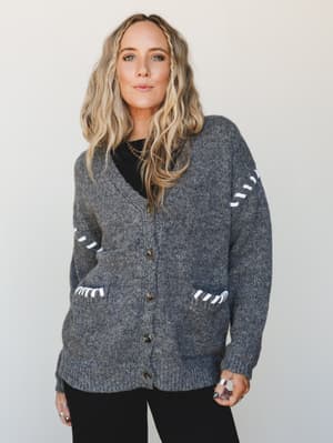 A chunky charcoal knit cardigan with white whip-stitch detailing along the sleeves and patch pockets, styled over a black top with silver rings and a chain necklace — the kind of cardigan you reach for every single weekend.