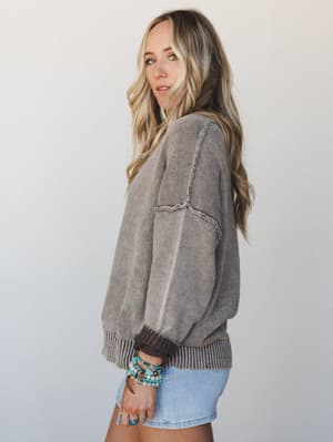 A relaxed knit sweater in washed brown with exposed seam detailing and balloon sleeves, styled with a denim mini and stacked turquoise bracelets — the kind of top that looks equally good on a coffee run or a slow Sunday.