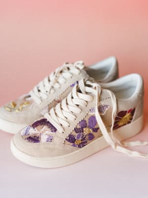 A pair of cream low-top sneakers covered in richly embroidered florals — purple, burgundy, and gold blooms across a natural canvas upper — shot against a soft blush pink background with a clean, close-in editorial feel.