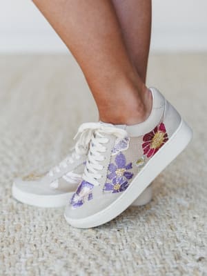 A close-up of floral embroidered sneakers on a model's feet, shot against a neutral interior backdrop — the kind of shoes that make a casual outfit feel like it had a whole plan.