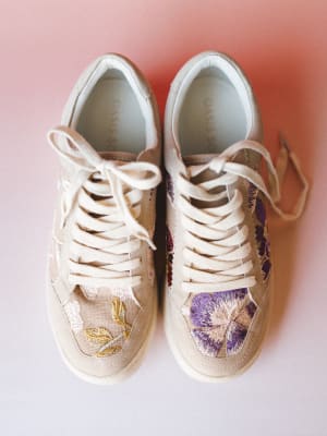 A pair of cream low-top sneakers covered in floral embroidery — lavender blooms on one toe, gold leafwork on the other — shot overhead against a soft pink gradient background.