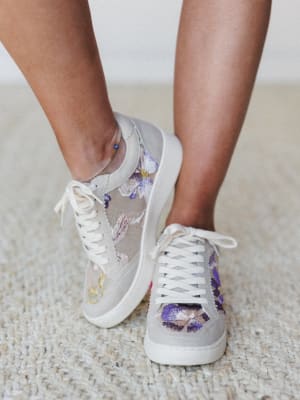 A close-up of floral embroidered sneakers on a model, showing off intricate pansy and botanical embroidery on a mesh and suede base — the kind of shoe that makes a plain outfit feel intentional.