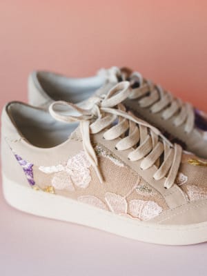 A close-up pair of lace-up sneakers in a warm taupe-cream tone with delicate floral embroidery across the side panel — blush pink blooms, soft purple flowers, and gold thread details against a dusty pink studio backdrop.