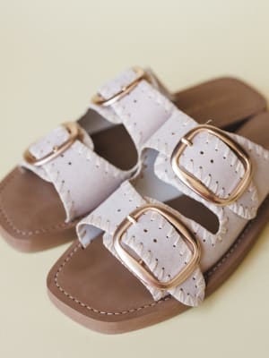 A pair of double-strap slide sandals in bone with oversized gold buckles, whipstitch edging, and a warm tan footbed — the kind of sandal that looks equally at home on a boardwalk or a patio brunch.