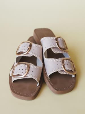 A pair of double-strap slide sandals in soft bone with oversized gold buckles, whipstitch detailing, and a warm brown footbed — shot on a muted sage background with that quiet, unhurried feel.