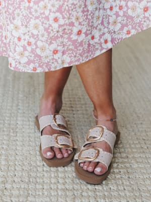 A close-up of double-buckle slide sandals in bone with gold hardware, paired with a flowy pink floral midi skirt — the kind of effortless warm-weather combo that looks like it wandered off a weekend farmers market.