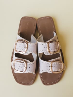 A pair of double-strap slide sandals in bone white laid flat overhead, featuring oversized gold buckles, perforated leather straps with whipstitch trim, and a warm brown footbed — the kind of sandal that goes with everything from a sundress to wide-l