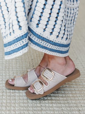 A close-up of wide-leg crochet pants in white and blue paired with bone slide sandals featuring oversized gold buckles — the kind of detail shot that makes you want to book a trip somewhere warm.