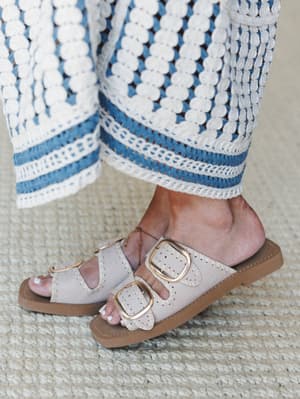 A close-up of wide-leg crochet pants in white and blue paired with bone slide sandals featuring oversized gold buckles — the kind of detail shot that makes you want to book a trip somewhere warm.