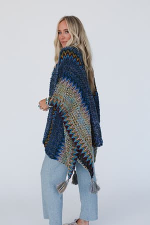 A woman with long blonde hair wears the Desert Dreams Crochet Ruana - Cobalt, a textured knit poncho with zigzag patterns and tassels, paired with light blue pants, standing against a plain white background.