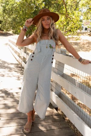 A woman in High Hopes Denim Overalls - Denim Stripe, a wide-brimmed brown hat, and sandals leans against a white fence on a sunny day, with trees and a dirt path in the background.