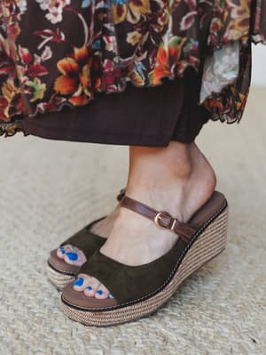 A close-up detail shot of a moss green suede platform wedge sandal with a tan leather ankle strap and woven jute-style platform, styled against a floral maxi skirt in deep brown and rust — earthy, grounded, and just a little unexpected with that pop