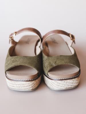 A pair of olive suede platform sandals shot straight-on against a soft blush-grey studio backdrop — the woven espadrille sole and cognac leather ankle strap give them that easy, well-traveled feel.